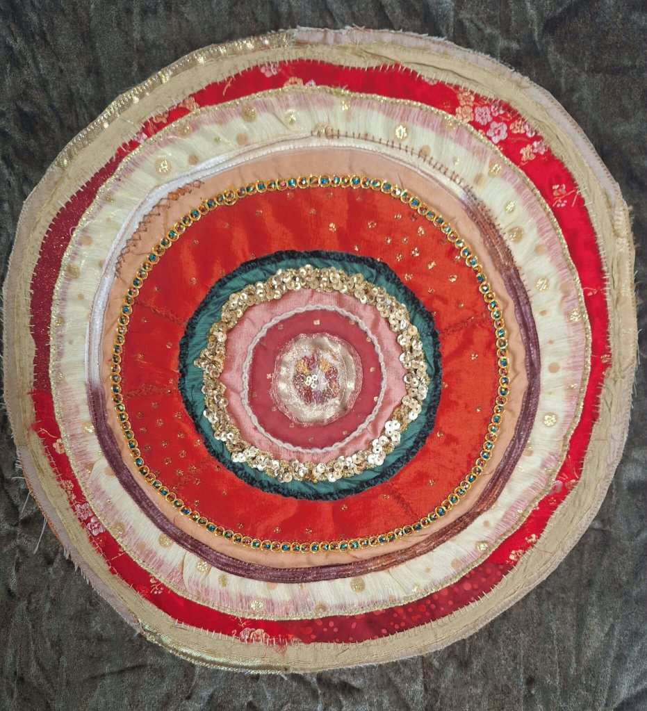 Red, cream, green and gold fabric mandala, with embroidery, sequins and ribbons attached.