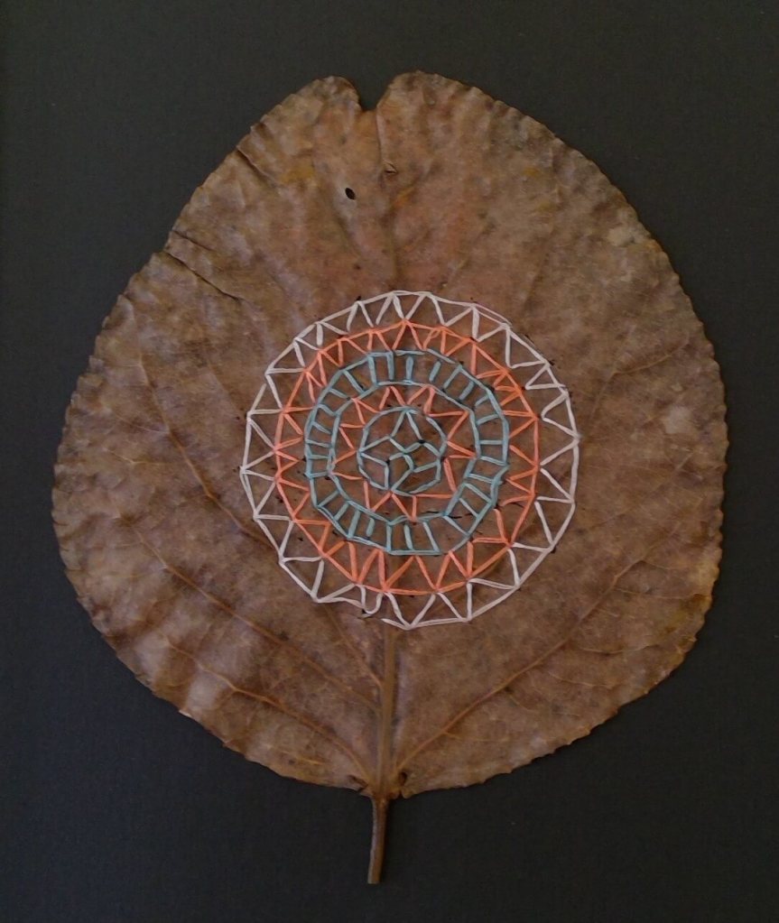 A real leaf hand embroidered with a geometric mandala pattern using white, orange and blue thread.