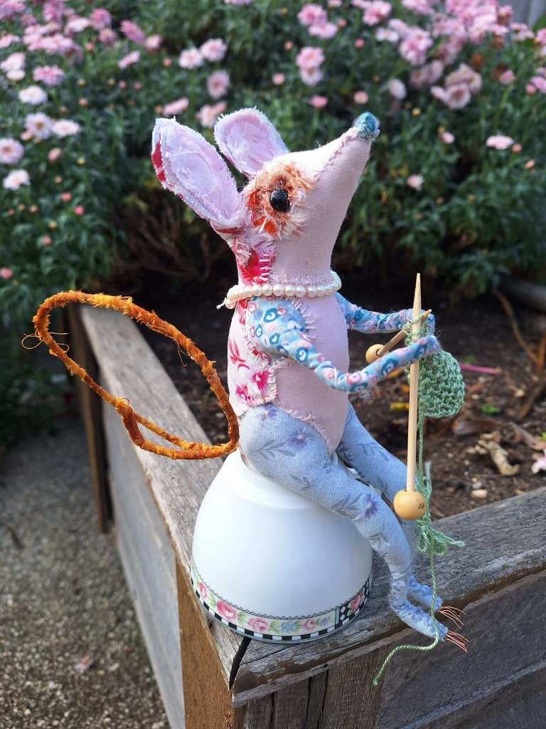 Textile mouse sculpture made from colourful fabrics. Knitting with tiny wooden knitting needles, sat on a china teacup in among the garden flowers.