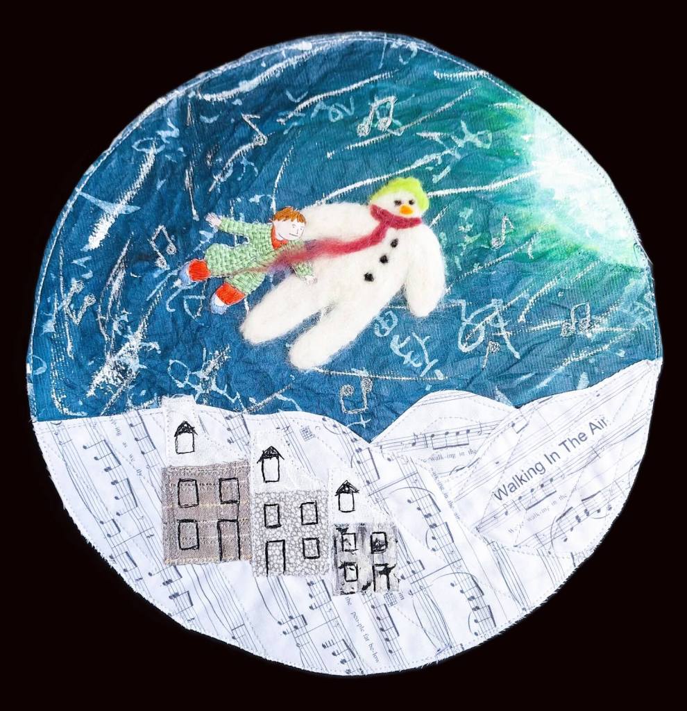 Circular mixed media artwork using paper, fabric, paint, stitching. Printed sheet music for snow covered hills, textile house, painted sky and 3d embroidered snowman and boy flying over the village below