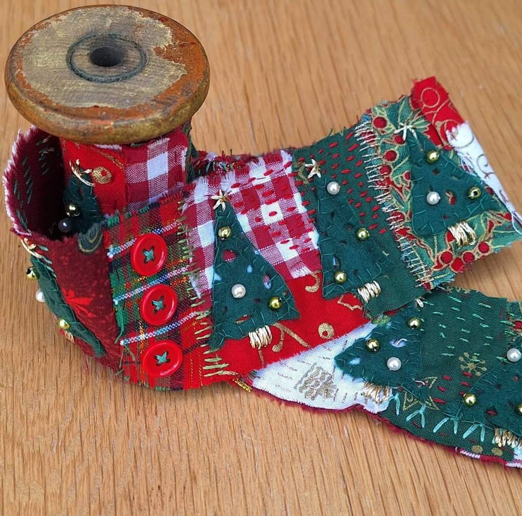Textile scroll with Christmas theme using red, green, gold and white fabrics, beads buttons and embroidery stitched. Displayed on a wooden spool.